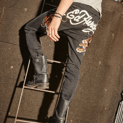 Model wearing black graphic jeans and black boots, seated on a rooftop ladder in an urban setting.