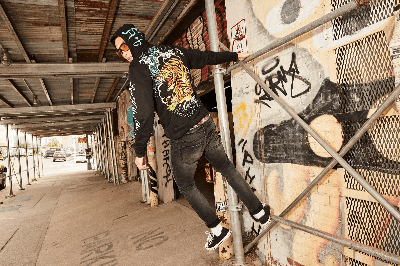 Man in a black Ed Hardy tiger graphic hoodie posing on scaffolding beside a graffiti-covered wall.