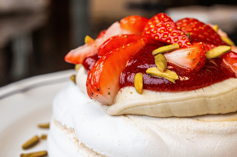 A macro shot of a pavlova’s topping, featuring glistening strawberry slices drizzled with red berry sauce and sprinkled with slivered pistachios.