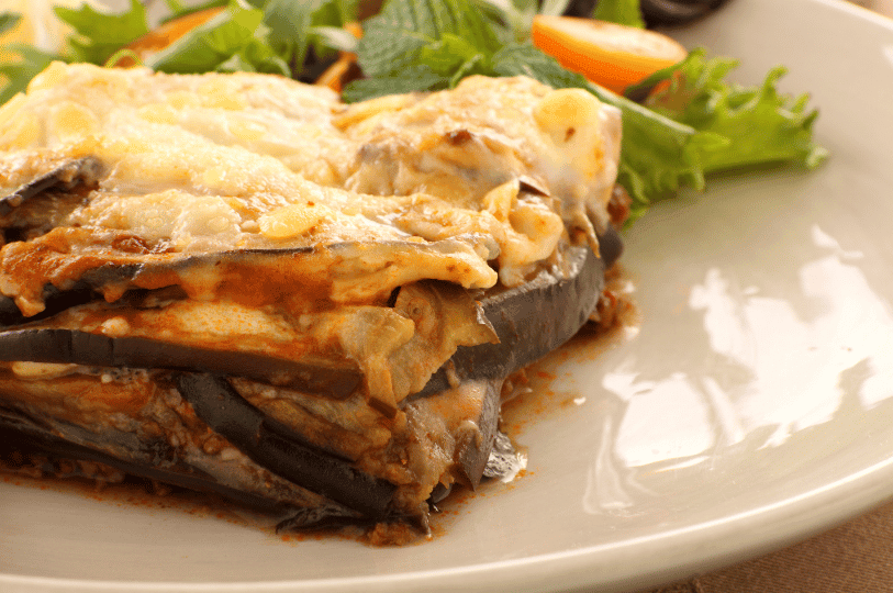 A close-up of Greek moussaka with golden melted cheese and creamy béchamel over layered eggplant and meat, garnished with mint, lettuce, and cherry tomato—highlighting its rich texture and vibrant Mediterranean presentation.