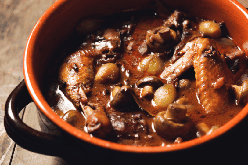 A close-up of Coq au Vin in a rustic red ceramic pot, showing tender chicken wings and whole mushrooms coated in a thick, glossy dark red wine reduction.