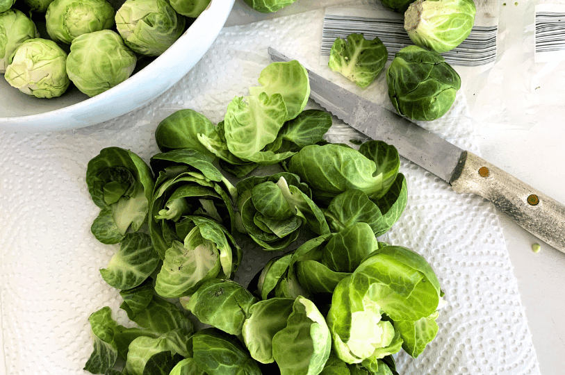 Raw Brussels sprout leaves and whole sprouts on a white paper towel next to a serrated kitchen knife, showing the prep process.