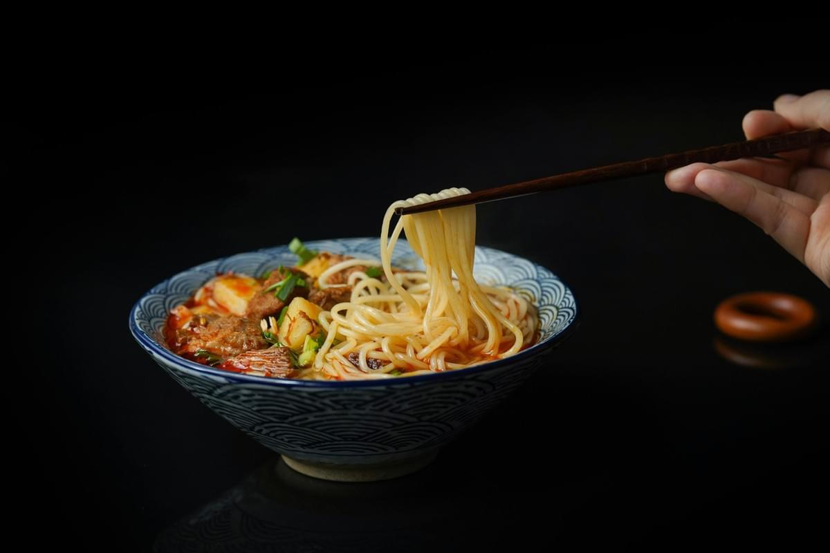 aAgainst a stark black background, a hand uses wooden chopsticks to lift thin noodles from a blue and white patterned ceramic bowl. The spicy, reddish broth is hearty, filled with chunks of braised meat and garnished with fresh green onions.
