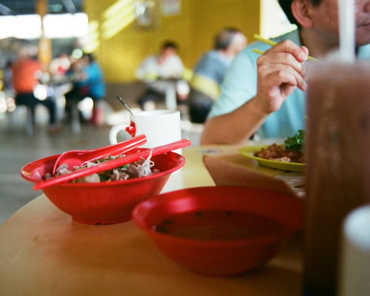 A bowl of noodles served on a table at a hawker centre, with blurred diners and food stalls in the background, showing a typical local dining scene.