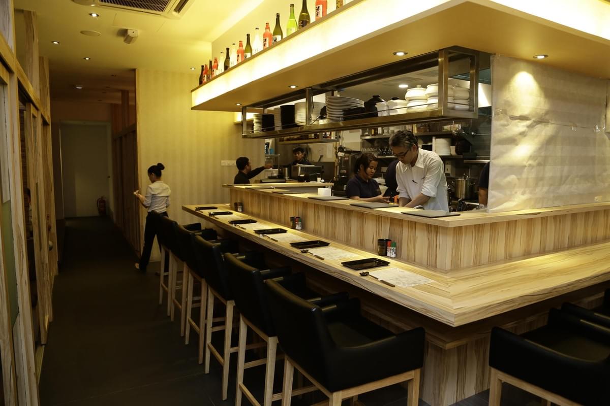 The image shows the interior of a modern Japanese restaurant featuring a light-colored wooden sushi counter lined with black bar stools. Several staff members are visible behind the counter and in the hallway, working under warm, ambient lighting.