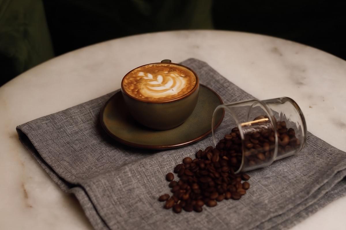 A cup of latte with leaf latte art on a saucer next to a glass of scattered roasted coffee beans on a grey napkin.