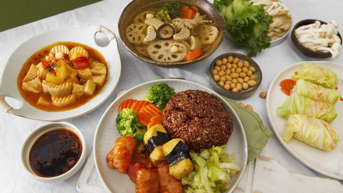 This image showcases a vibrant spread of vegetarian dishes, including a central plate with red rice, steamed vegetables, and seaweed-wrapped tofu. The surrounding table is filled with various side dishes, such as a lotus root soup, sweet and sour stir-fried vegetables, and delicate cabbage rolls, all arranged on a white cloth.