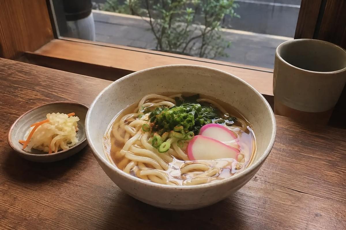 A high-angle, close-up shot of a steaming bowl of traditional Japanese udon noodle soup served in a speckled light-cream ceramic bowl on a rustic dark wooden counter next to a window. The thick, chewy noodles float in a clear, golden dashi broth, topped with chopped scallions, a dash of red shichimi pepper, dark green seaweed, and two slices of pink-rimmed kamaboko fish cake, accompanied by a small side dish of pickled vegetables to the left and a neutral-toned ceramic tea cup sitting to the right against the backdrop of an outdoor garden view.
