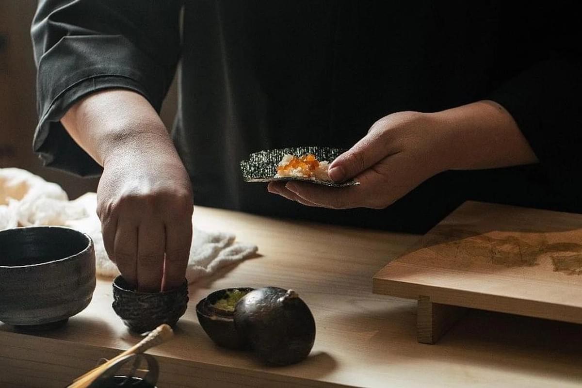 A chef meticulously prepares a hand-rolled sushi course, placing bright orange roe onto a crisp sheet of nori. The scene captures an intimate moment of culinary craftsmanship with dark, textured ceramics and a wooden workspace.
