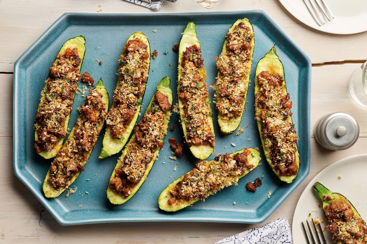A blue platter holding several halves of zucchini (zucchini boats) stuffed with a savory meat and breadcrumb mixture, garnished with herbs.