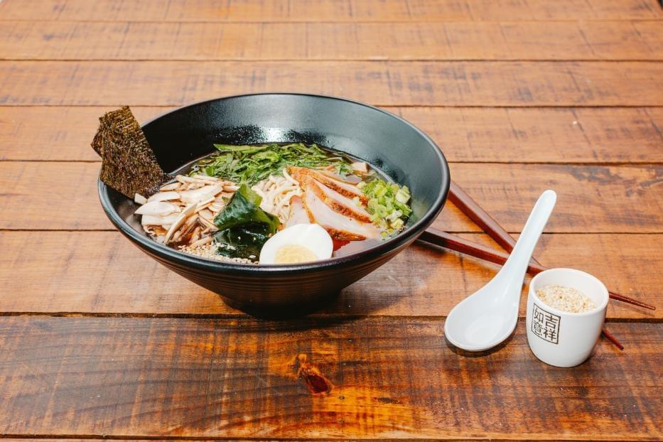 A black bowl filled with a colorful ramen noodle soup sits on a warm wooden tabletop. The dish is meticulously arranged with sliced pork, a soft-boiled egg, mushrooms, greens, seaweed, and scallions, accompanied by a white spoon, chopsticks, and a small cup of sesame seeds.