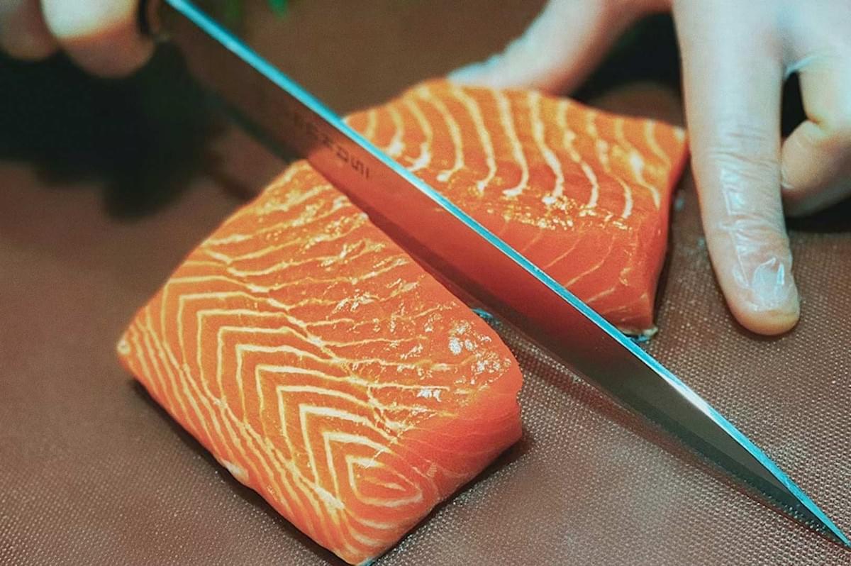 Gloved chef slicing two raw salmon fillets with a sharp knife, highlighting precise cuts and bright orange fish texture.