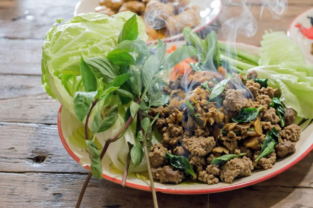 A close-up of a steaming plate of hot Laotian Larb featuring cooked minced meat mixed with herbs, served alongside fresh basil leaves and a large wedge of cabbage on a wooden table