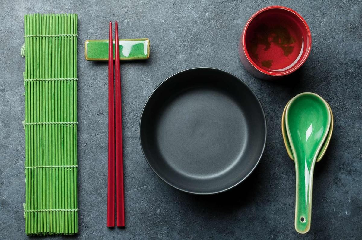 Sushi-making tools flatlay: bamboo rolling mat, red chopsticks on ceramic rest, black mixing bowl, and green ceramic spoon on dark textured surface.