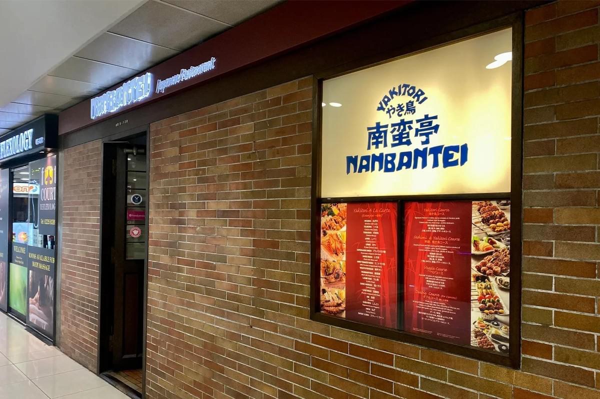 The storefront of Nanbantei Japanese Restaurant features a brick exterior and a lit sign displaying the restaurant's name in English and Japanese. A large menu board is displayed next to the entrance, showcasing various yakitori courses and dishes.