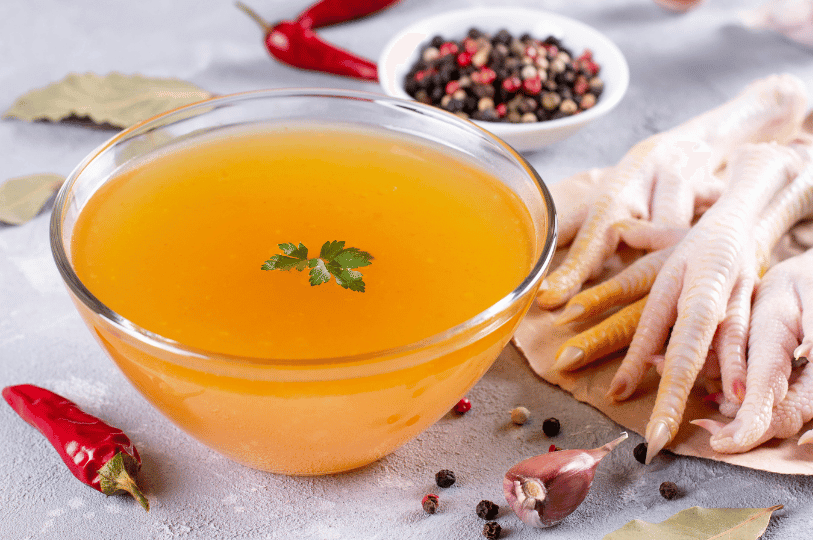 A glass bowl filled with golden chicken stock surrounded by raw chicken feet, dried red chilies, peppercorns, and garlic cloves.