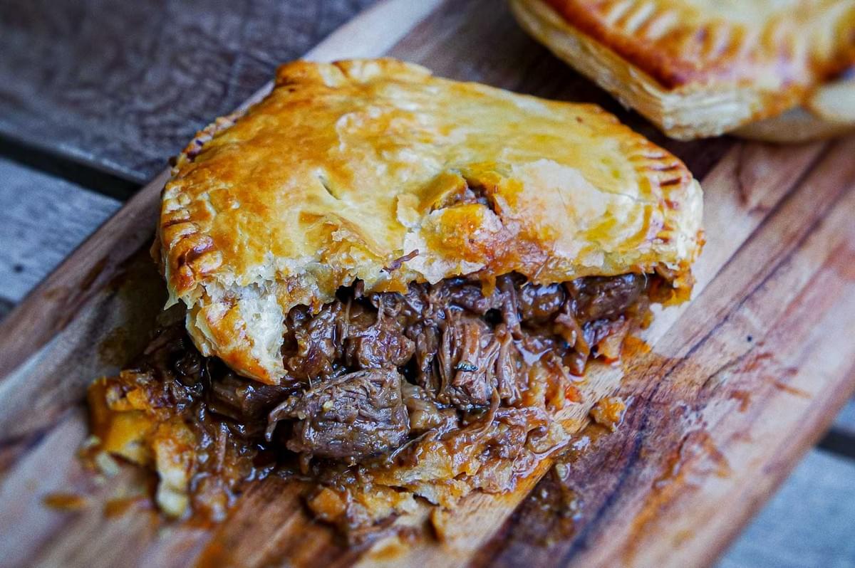 Australian meat pie with golden flaky crust, crimped edges, and shredded beef filling in rich gravy, served on rustic wooden board.