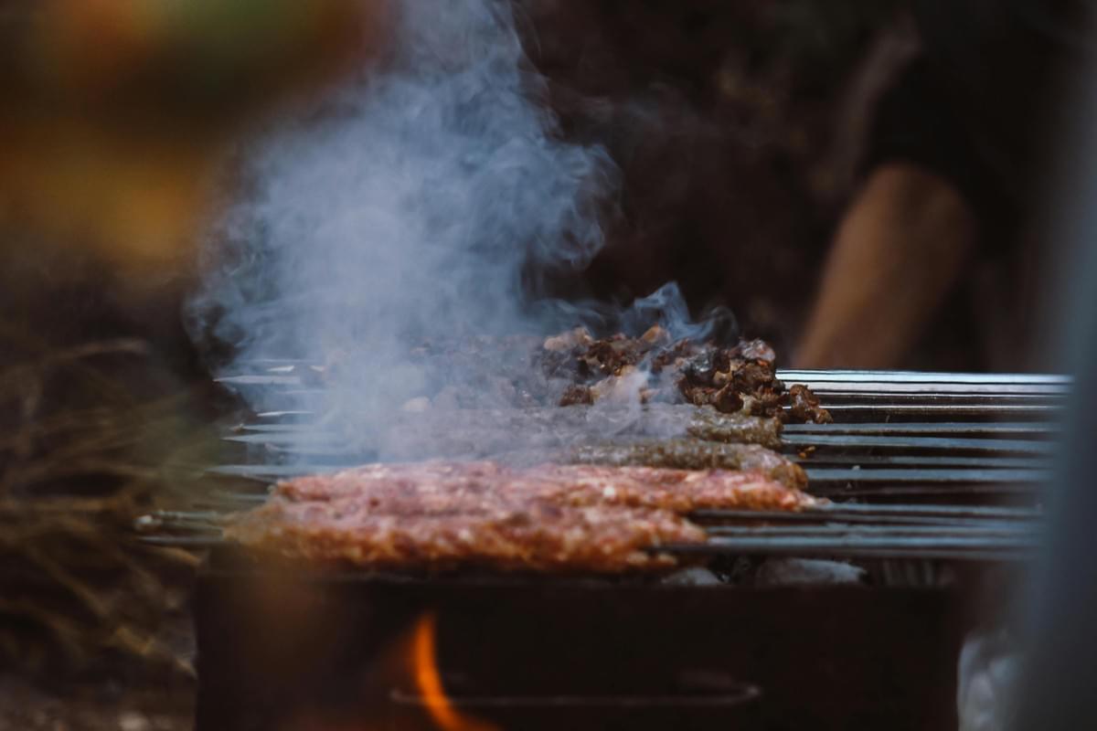 Metal skewers laden with ground meat are grilling over an open fire, enveloped in thick, rising smoke. Visible flames lick at the bottom of the grill, cooking the kebabs in what appears to be a rustic, outdoor setting.