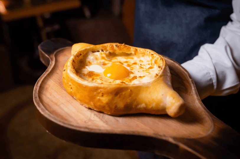 server's hands holding a rustic wooden board featuring a deep-dish Khachapuri. The bread has high, thick crusts and a deep well of melted cheese topped with a sunny egg yolk, captured in warm, atmospheric restaurant lighting.