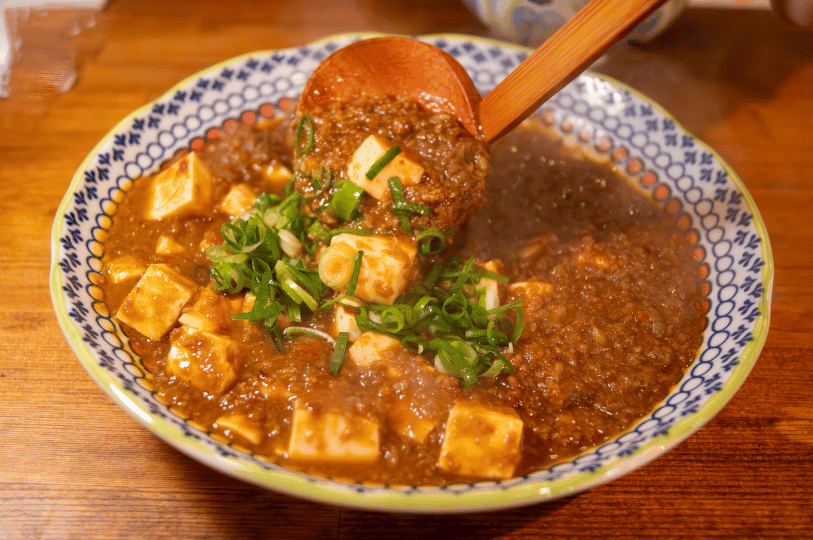 A wooden ladle scooping up a serving of Mapo Tofu from a patterned bowl, highlighting the glossy texture of the spicy sauce and the contrast of green scallions.