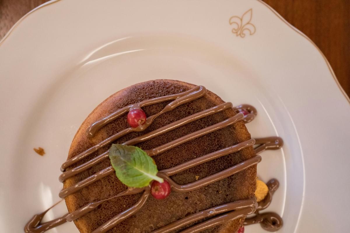 An overhead shot shows a small, round sponge cake on a white plate with a gold fleur-de-lis emblem. The cake is decorated with a drizzle of chocolate sauce, a fresh green mint leaf, and a few small red berries.