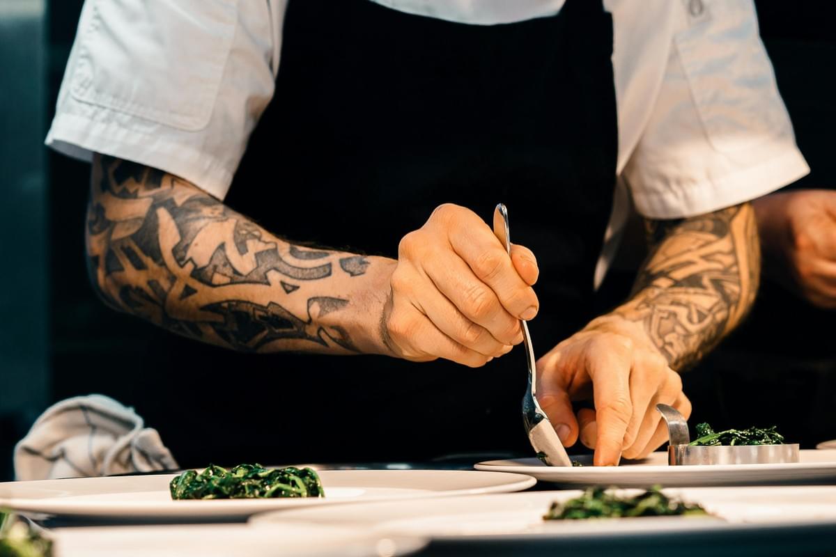 A close-up captures a chef with heavily tattooed arms meticulously plating food in a kitchen. Wearing a black apron and white chef's coat, they use a spoon and a metal ring mold to precisely arrange bright green greens onto white plates.