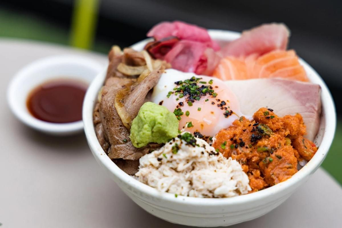 A white ceramic bowl is filled with a colorful surf-and-turf Japanese rice bowl topped with thin-sliced cooked beef, various cuts of fresh sashimi, crab meat, spicy tuna, pickled ginger, and a soft-poached egg. A small dollop of wasabi sits near the center, with a small dish of soy sauce placed in the blurred background.