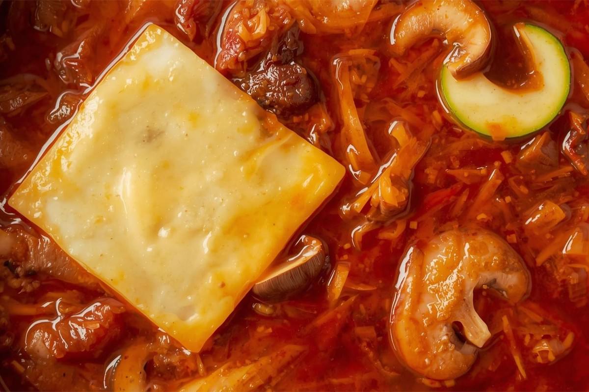 A dramatic macro shot of a single square of melting cheese floating in a rich, red broth with slices of mushroom and zucchini, suggesting a variation like Budae Jjigae (Army Stew).
