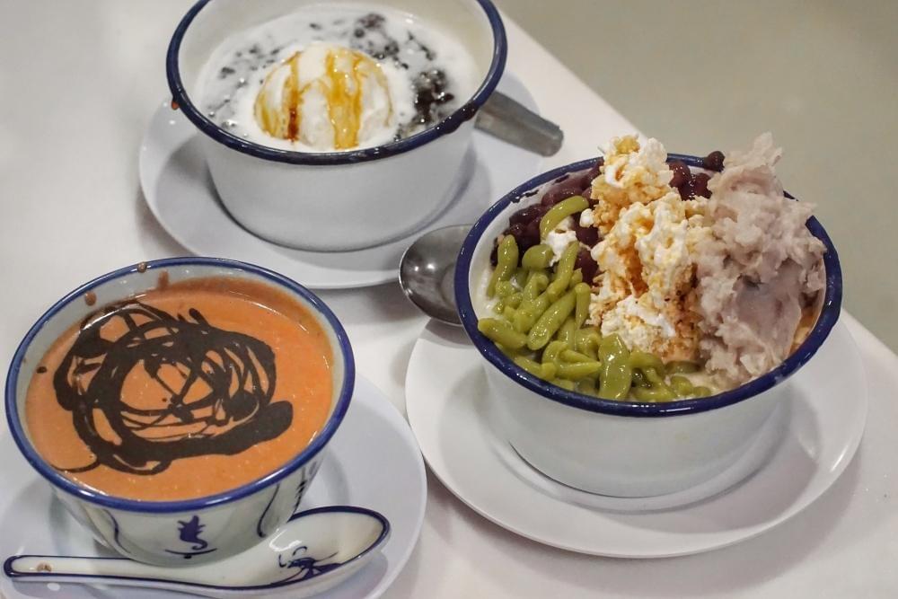 Three bowls of traditional Southeast Asian desserts are arranged on a table, featuring colorful ingredients like green pandan jelly, red beans, and taro paste. The presentation includes a bowl of black glutinous rice topped with ice cream and a warm orange-hued soup drizzled with dark syrup.
