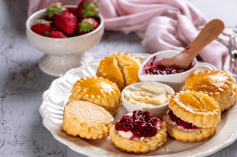 An elegant teatime spread featuring several scones on a scalloped plate, served with small bowls of clotted cream, jam, and a side bowl of fresh strawberries.