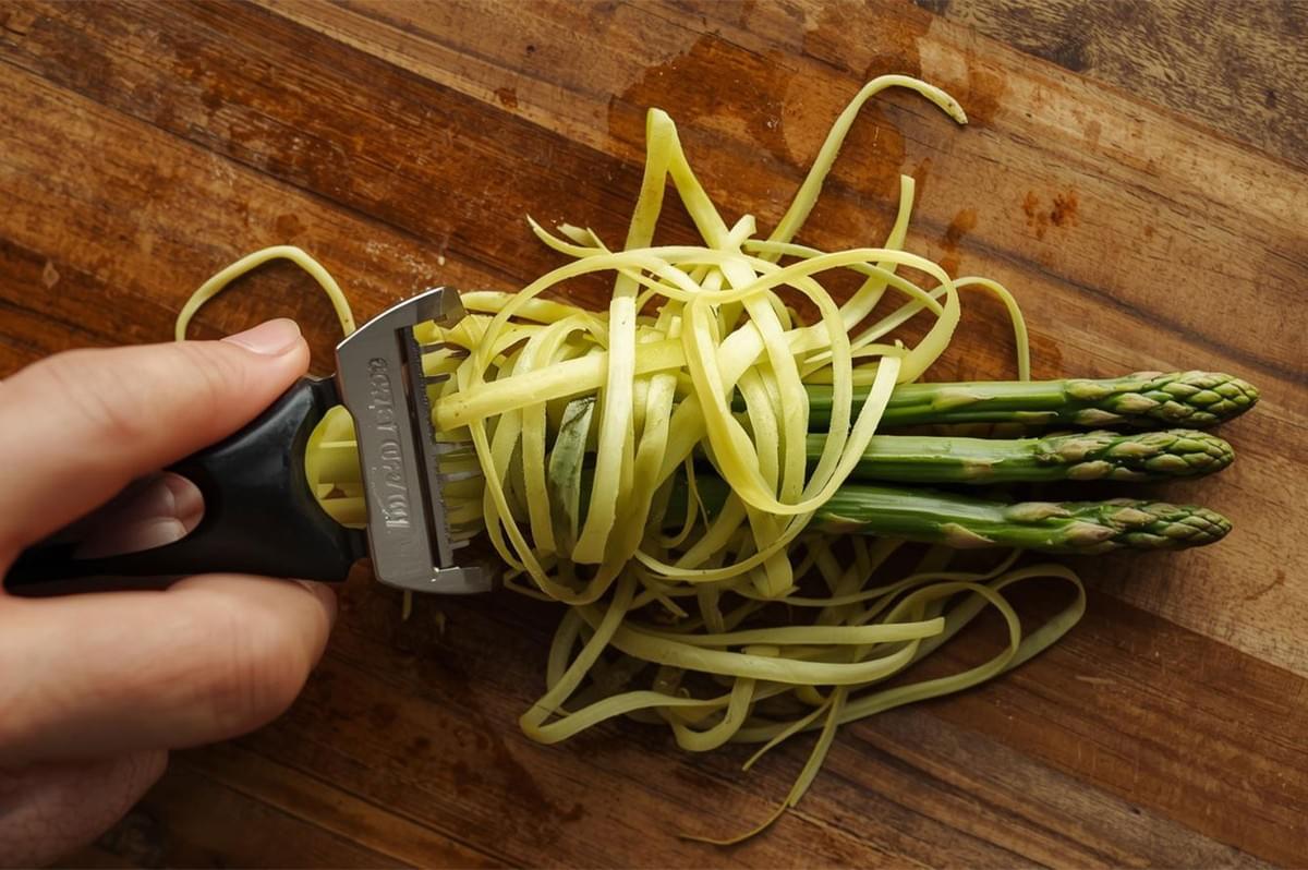 An overhead shot of a hand using a vegetable peeler (specifically a multi-blade shaver) to create delicate, thin, raw asparagus ribbons on a wooden cutting board.