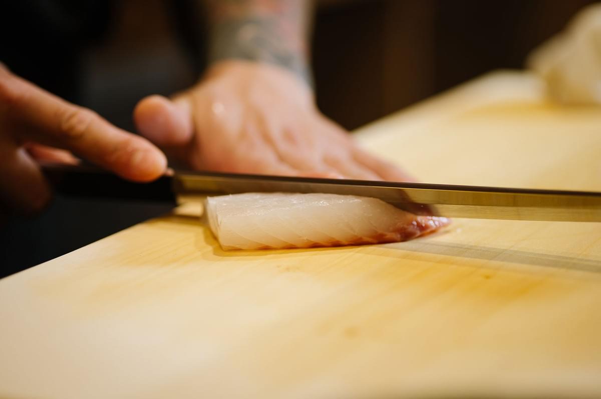 Close-up of a chef's hands using a sharp Japanese knife to precision-slice a fresh piece of white fish sashimi on a wooden cutting board.