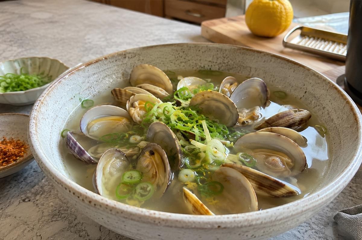 A large, speckled ceramic bowl sits on a light gray countertop, filled with steamed clams resting in a light, semi-translucent broth. The opened clam shells reveal tender meat inside, and the dish is heavily garnished in the center with a mound of finely sliced green scallions and bright yellow yuzu or lemon zest. In the background, a wooden cutting board holds a whole yellow citrus fruit next to a microplane grater, while small side bowls containing extra sliced scallions and red shichimi torgarashi spice blend sit to the left of the main dish.