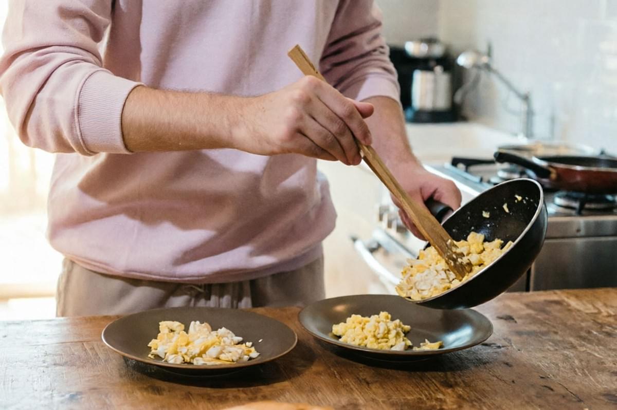 Person serving scrambled eggs from pan onto plates on wooden table