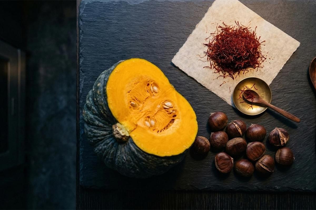 Flat lay of raw ingredients including a halved kabocha squash, a pile of red saffron threads, and whole chestnuts on a dark slate board.