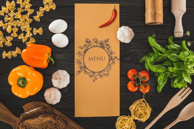 Flat lay of a mustard-yellow menu card centered on a dark wooden surface, surrounded by raw culinary ingredients including fresh basil, cherry tomatoes, yellow bell peppers, garlic cloves, eggs, and scattered dry pasta.