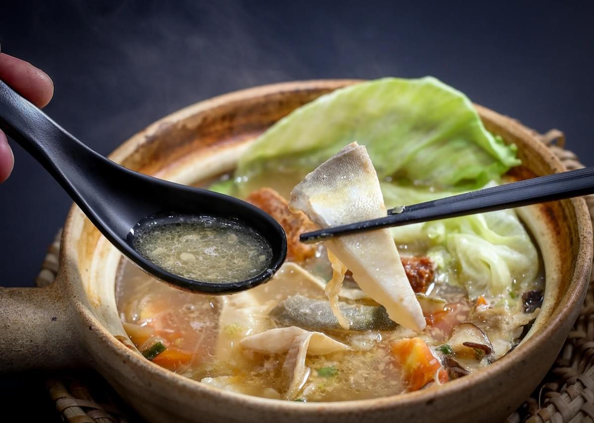 A steaming clay pot filled with hot fish soup, cabbage, and vegetables sits on a dark table. A hand holds a black spoon scooping broth, while chopsticks lift a tender slice of pomfret fish above the bowl.