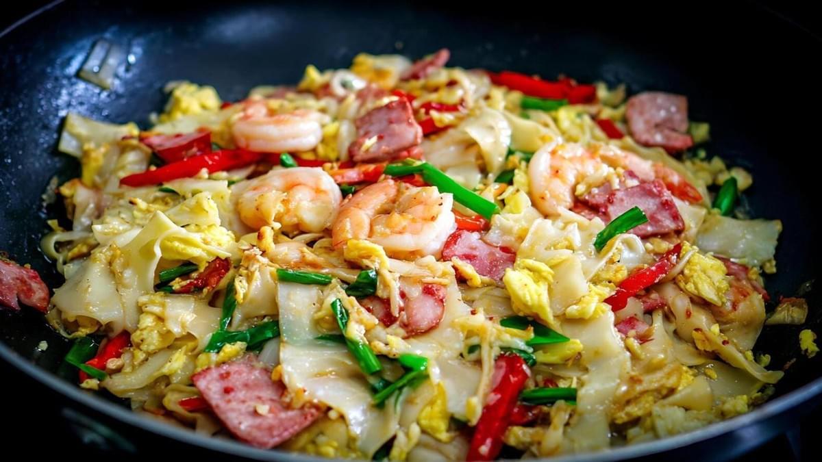  This close-up shot features a colorful stir-fried noodle dish being cooked in a black wok or pan. The wide, flat noodles are mixed with ingredients such as pink shrimp, pieces of red meat likely Chinese sausage or ham, scrambled egg, bright red chili slices, and chopped green onions.