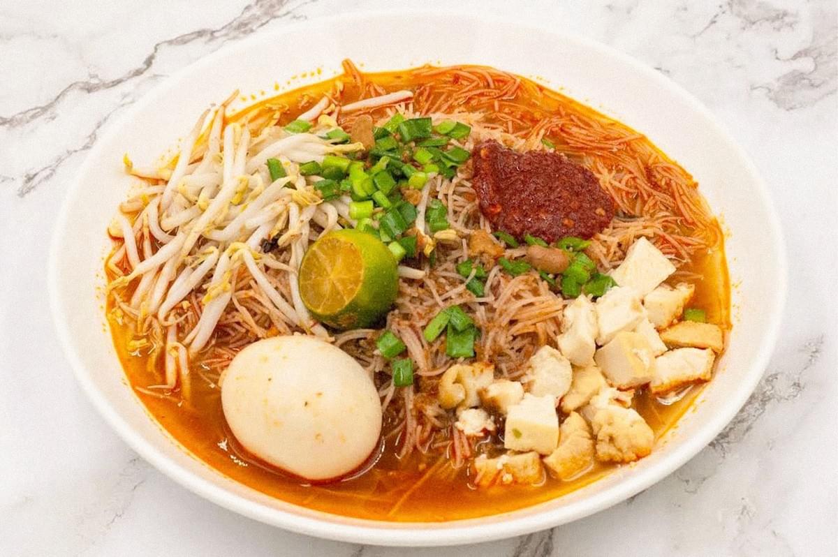 A bowl of mee rebus with thin rice vermicelli, bean sprouts, tofu, a boiled egg, chopped green onions, lime, and a piece of sambal.