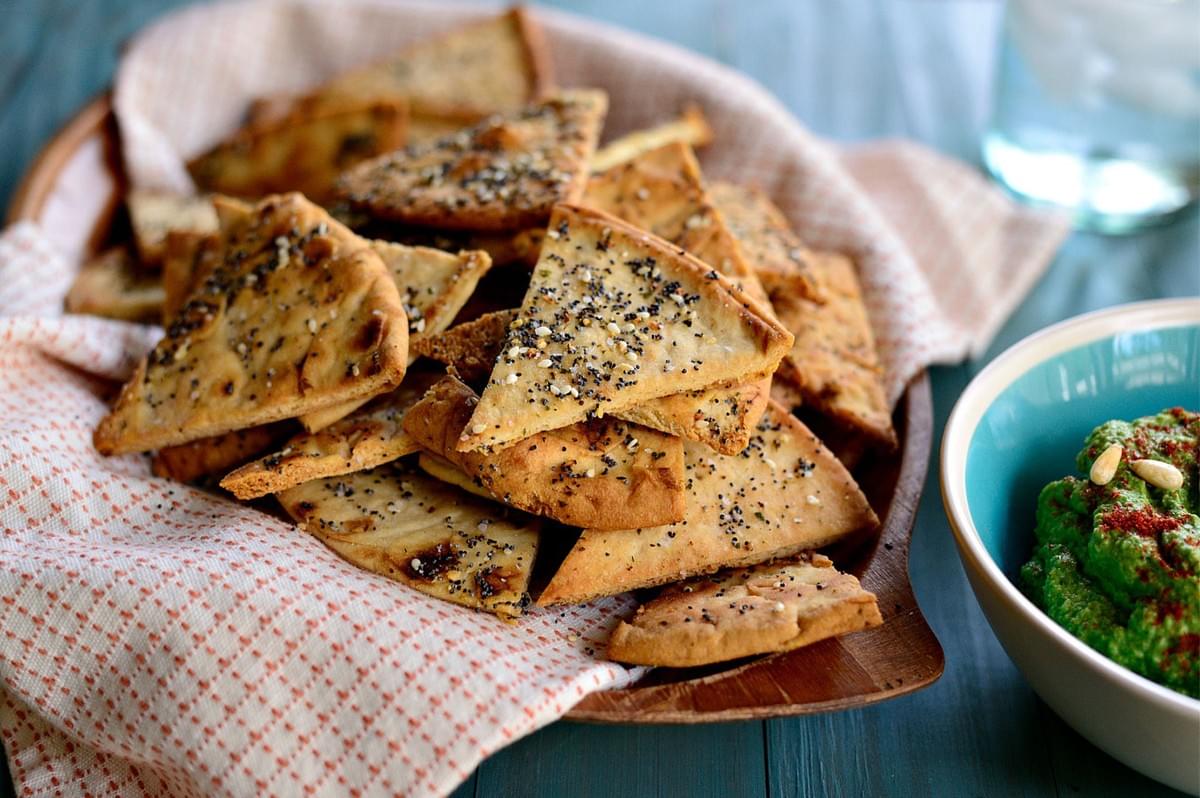 A wooden bowl lined with a patterned cloth, holding a large pile of homemade, triangular baked pita chips sprinkled with sesame seeds and za'atar seasoning, next to a small bowl of green hummus.