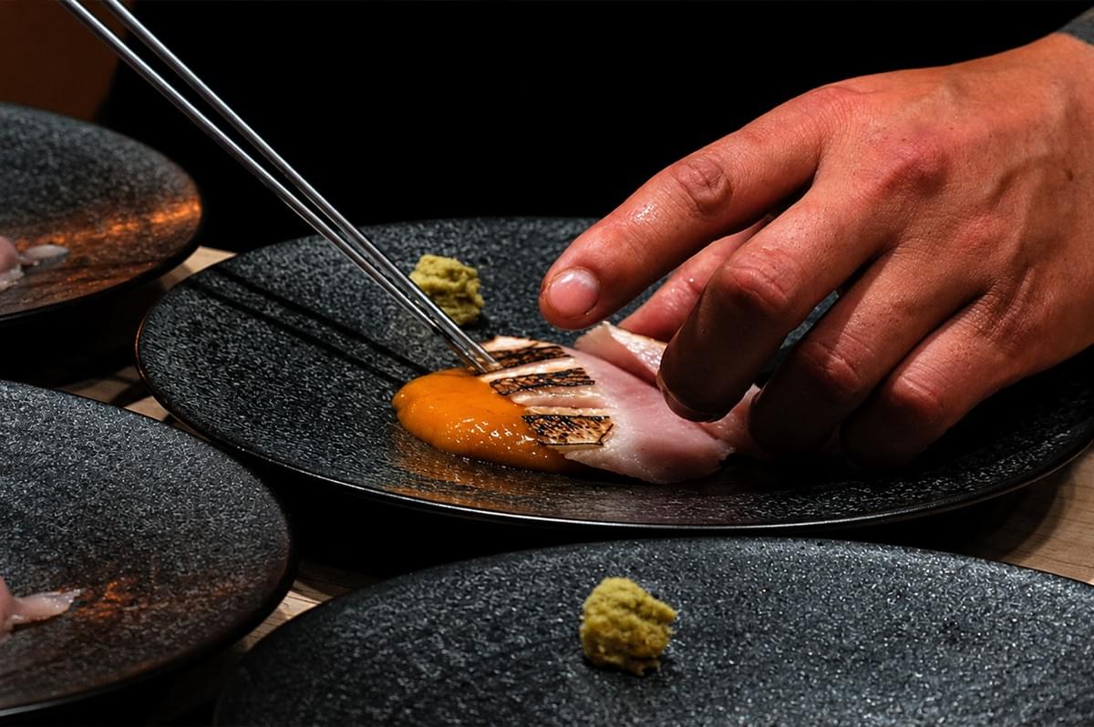 Features a close-up, high-angle view of a chef's hands meticulously plating an upscale dish on dark, textured ceramic plates. The chef uses slender metal culinary tweezers in one hand and gently guides a slice of lightly seared, pale pink fish or meat with the other, arranging it over a vibrant, smooth orange puree. Each plate includes a small, neat dollop of green wasabi or herb paste off to the side, and several identical plates are partially visible in the frame, suggesting a professional kitchen line or tasting menu assembly. The lighting is focused and dramatic, highlighting the texture of the chef's skin, the grill marks on the food, and the grainy surface of the plates against a dark background.