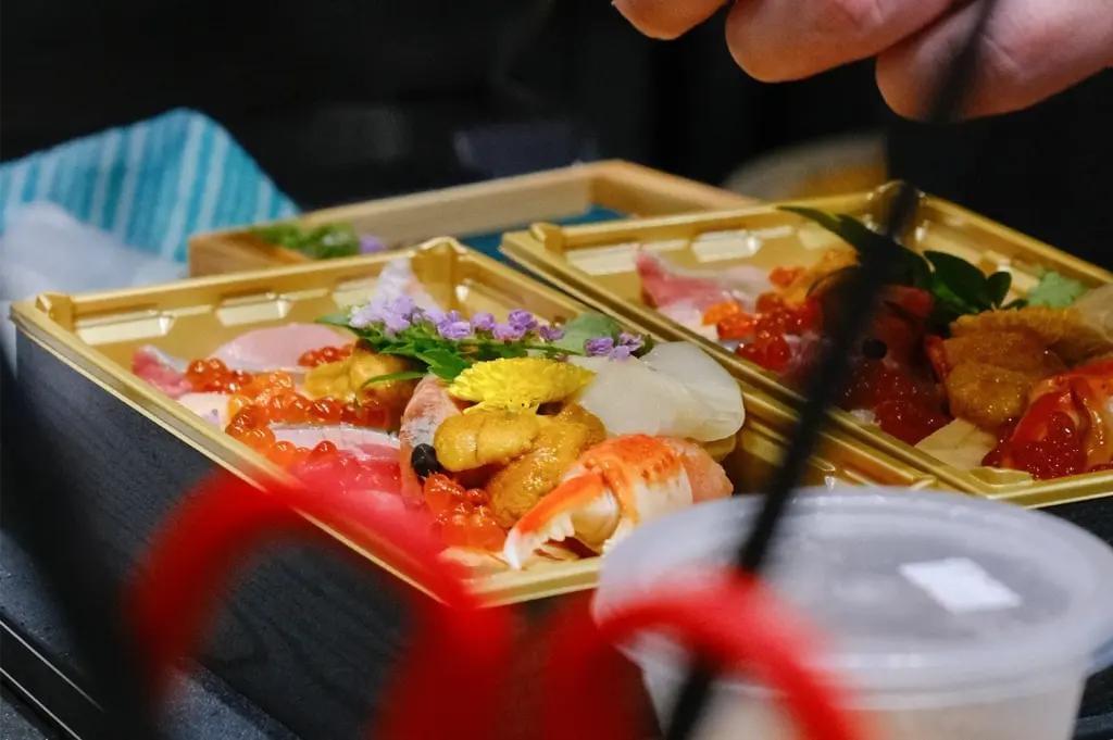 A close-up view of Japanese kaisen-don seafood bento boxes filled with fresh ingredients like sea urchin, salmon roe, crab claw, and raw fish. The colorful dishes are garnished with delicate edible flowers and served in golden containers.