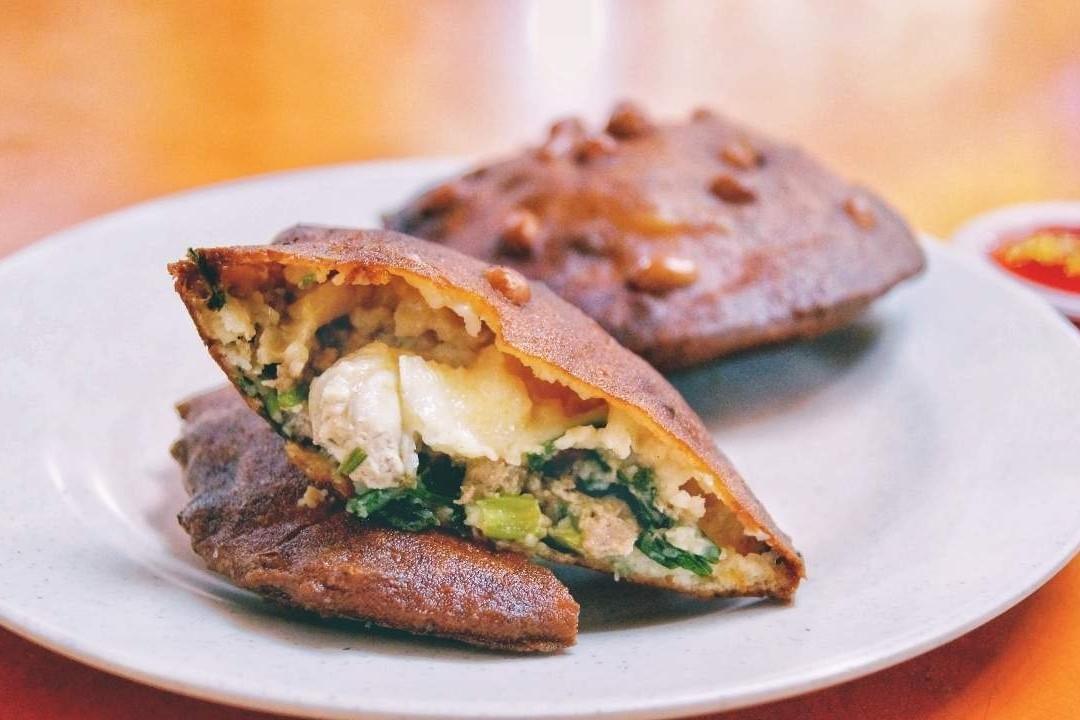 A plate holds two golden-brown, fried Fuzhou oyster cakes, with one sliced open to reveal a savory filling of oysters, meat, and greens. Topped with crunchy peanuts, these traditional snacks are served with a side of chili sauce on a bright orange table.