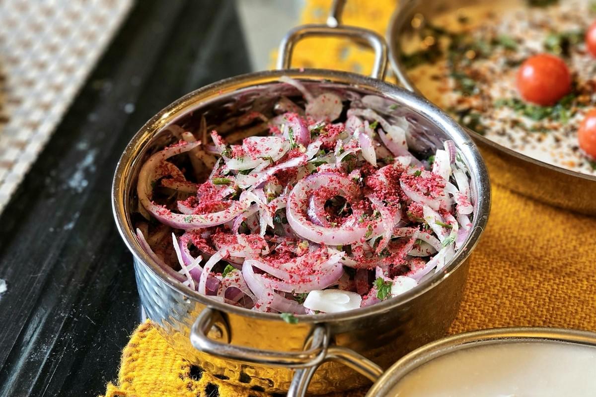 A small stainless steel bowl is filled with thinly sliced red onions mixed with fresh green herbs and generously dusted with a vibrant pink spice, likely sumac. The bowl sits on a yellow woven placemat next to another serving dish containing a creamy dip garnished with cherry tomatoes.