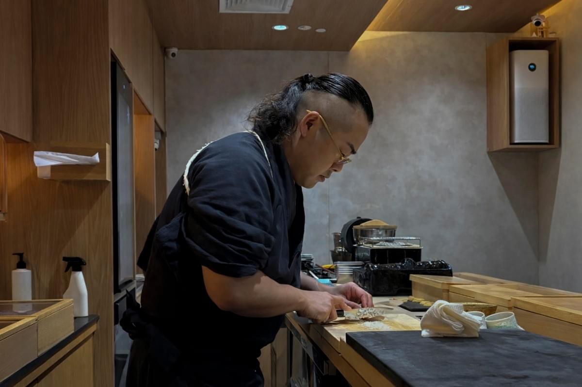 A side-profile shot of a focused sushi chef with a distinctive undercut hairstyle and a long ponytail. He wears thin-rimmed glasses and a dark traditional Japanese work tunic (samue). He is standing behind a clean, light-wood counter, skillfully using a long kitchen knife to prep ingredients on a wooden cutting board. The background shows a modern, minimalist kitchen with warm wood cabinetry, a grey textured wall, and an air purifier.