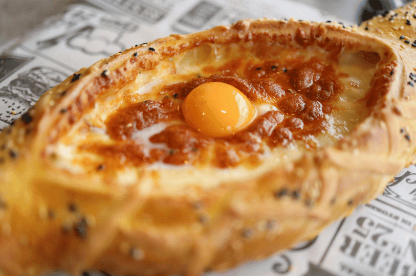 A macro shot focusing on the texture of a Khachapuri. The image shows the charred, bubbly cheese surface and a translucent egg yolk, with the bread crust sprinkled with black sesame seeds, resting on newsprint-style parchment paper.