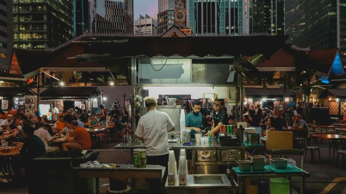 Set against a backdrop of towering modern skyscrapers, a bustling open-air food market comes alive at night with crowded tables of diners. In the foreground, busy workers attend to customers at a brightly lit beverage stall, highlighting the vibrant contrast between traditional street food culture and the urban city environment.