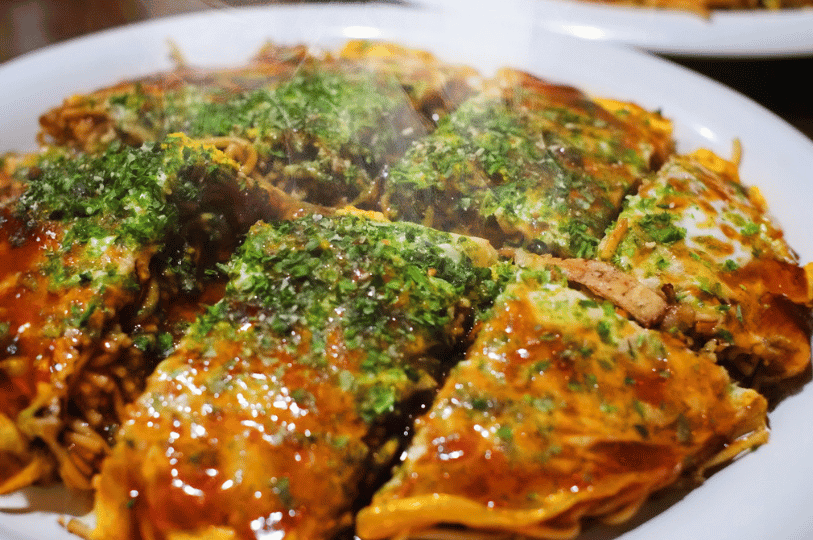 A steaming plate of okonomiyaki sliced into triangles, glistening with sauce and aonori, its rich, savory layers inviting a taste of Japan’s ultimate customizable pancake.