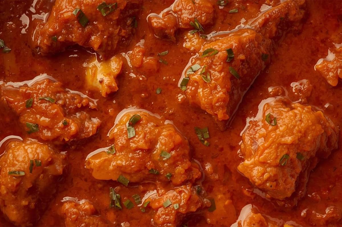 A close-up view of bright, fiery red Kashmiri Rogan Josh (likely lamb or chicken), featuring pieces of meat swimming in a rich, non-creamy, oil-glossed sauce, garnished with chopped chives.