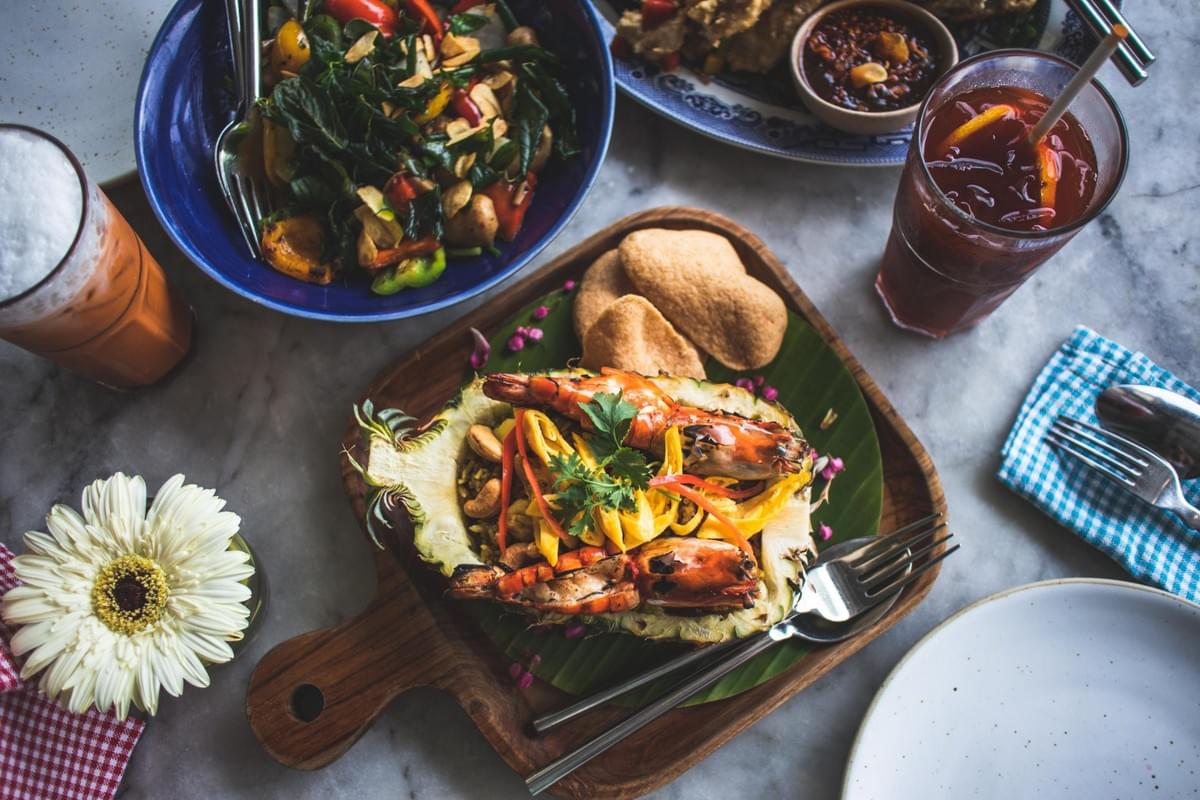 A flat-lay view of a table spread featuring pineapple fried rice served inside a carved pineapple boat topped with grilled prawns and cashews. Surrounding the main dish are a vibrant stir-fried vegetable bowl, prawn crackers, iced drinks, and a white daisy accent.